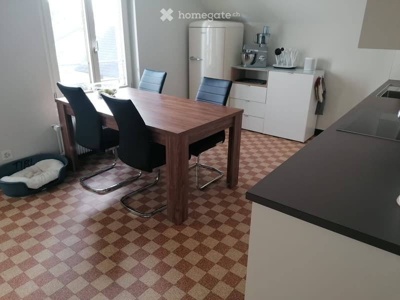 A modern kitchen and dining area with a wooden dining table, four chairs, a refrigerator, and a kitchen counter. The floor is checkered, and there is a dog bed in the corner.