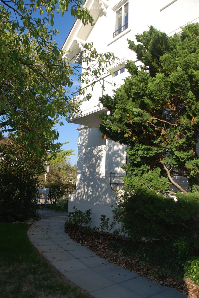 A white two-story house with a well-maintained garden, featuring a paved pathway leading to the entrance, surrounded by greenery and trees.