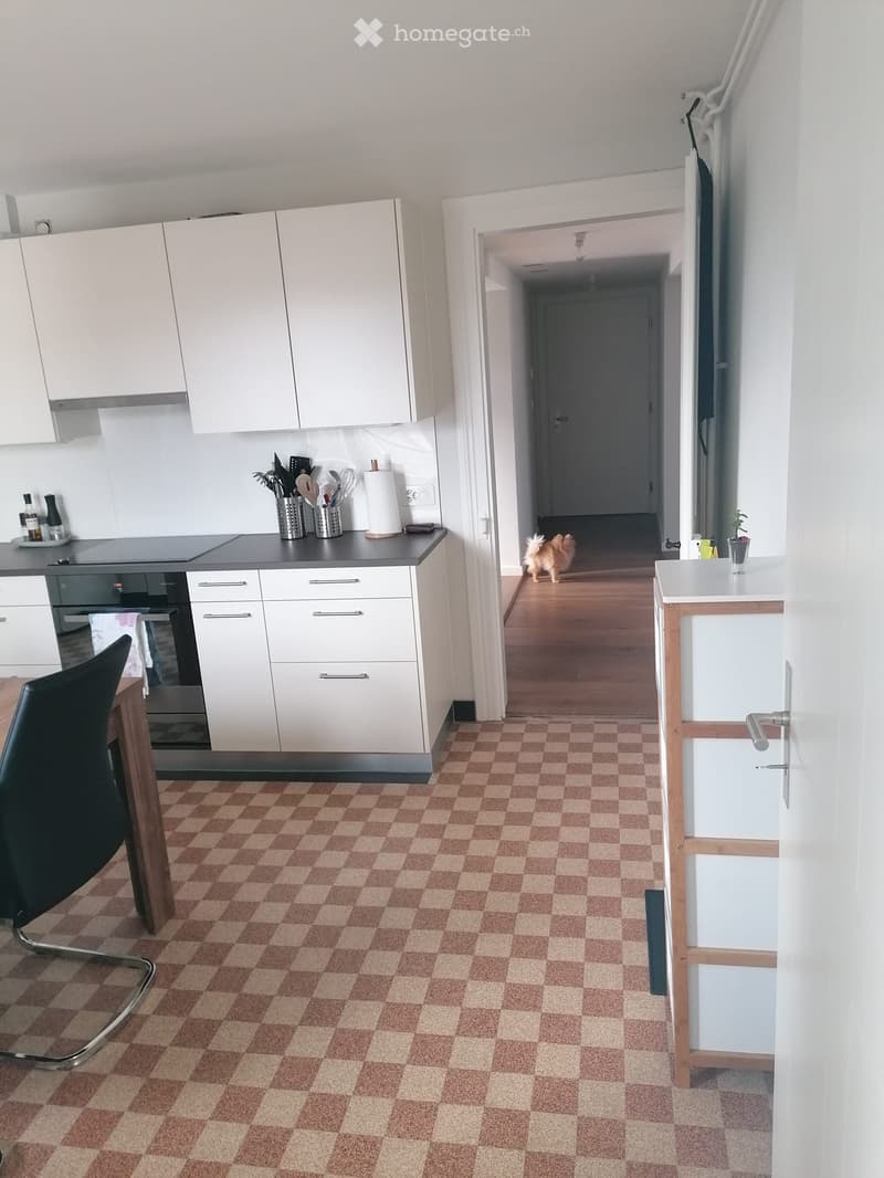 Modern kitchen with white cabinets, grey countertop, and checkered floor. Includes a chair, oven, sink, and various utensils on the counter.
