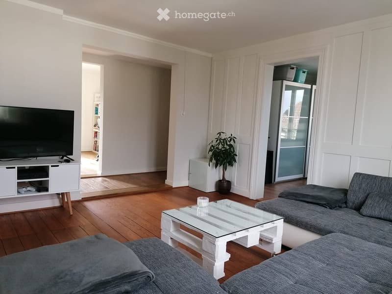 Modern living room with wooden flooring, grey sofa, glass coffee table, flat-screen TV on a white cabinet, and a potted plant near the doorway.
