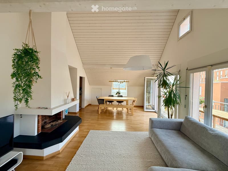 Modern living room with a grey sofa, coffee table, fireplace, dining area with a table and chairs, pendant light, and large windows leading to a balcony.