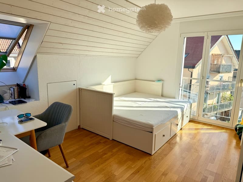 Bright, modern bedroom with wooden floors, a built-in bed, a desk with a chair, and a large window offering a view of the neighborhood.
