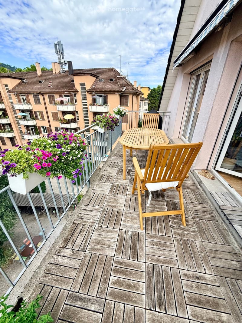 Balcony with wooden decking, a table, and a chair, overlooking a residential area with colorful flower boxes and neighboring buildings.