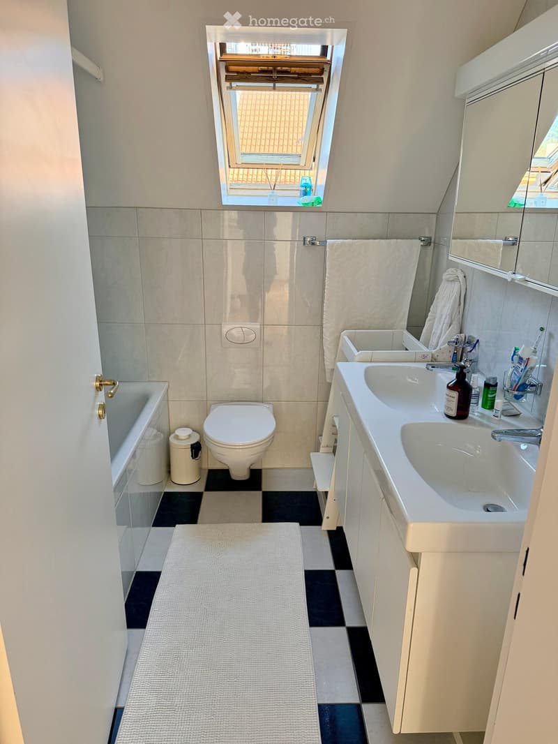 Modern bathroom with a bathtub, toilet, sink, and mirror. Features a white tiled wall, a window with blinds, and a white towel hanging on a rack.