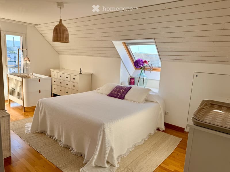 Cozy attic bedroom with wooden slat walls, skylight window, bed with white bedding and purple pillow, baby crib, nightstand with vase of flowers, and a wicker basket on the floor.