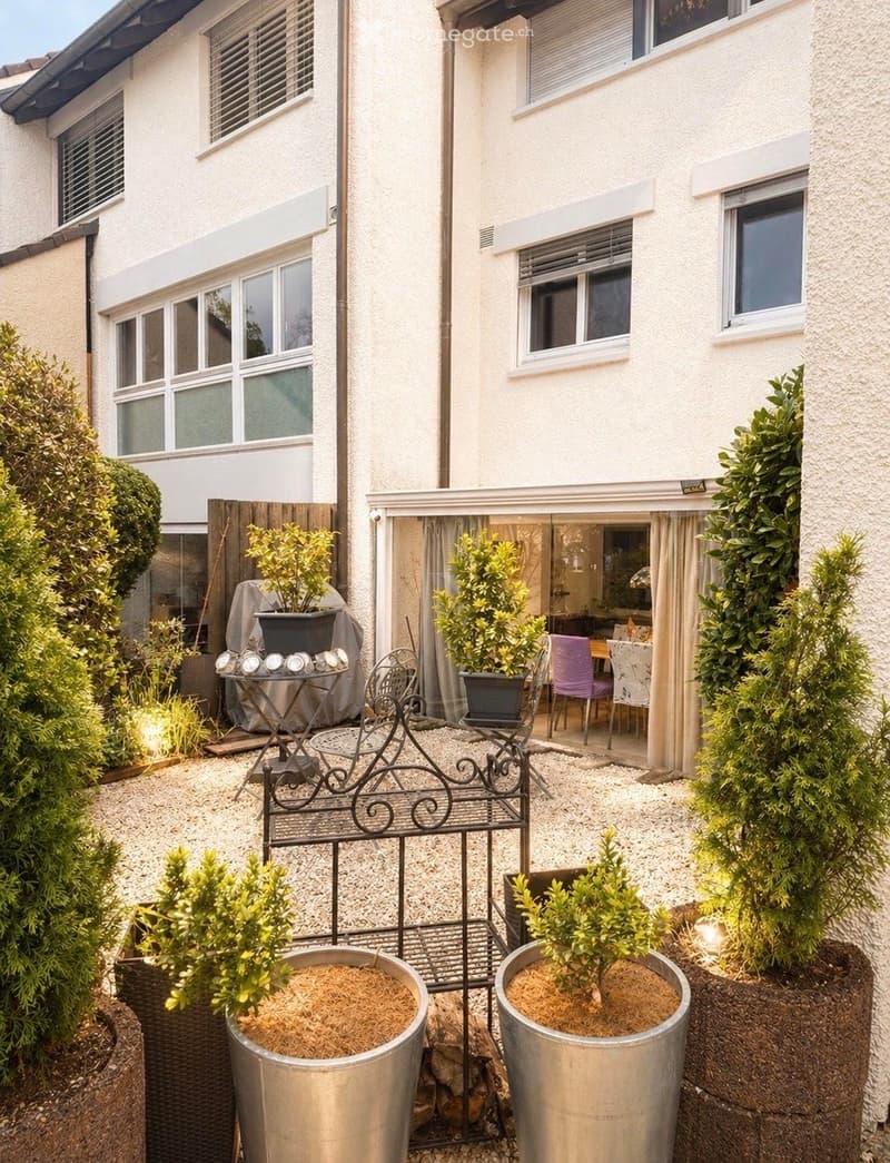 A small, well-maintained garden area with gravel flooring, several potted plants, a metal bench, and a small table with chairs. The garden is enclosed by a modern building with large windows and white walls.