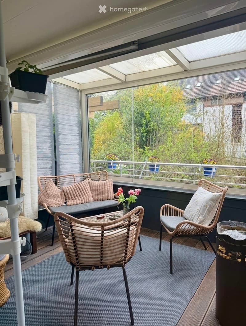 A modern, enclosed balcony with a sofa, two wicker chairs, a small table, and a vase with flowers. The space features a grey rug and a view of greenery outside.