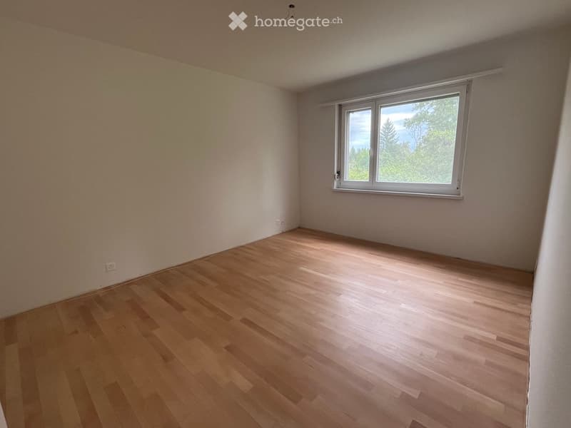 Empty room with wooden flooring and a large window showing a view of trees.