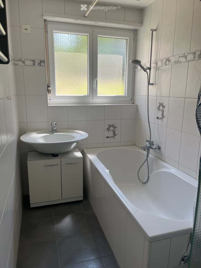 Modern bathroom with a bathtub, sink, and cabinet. The room features white tiles and a window with frosted glass.