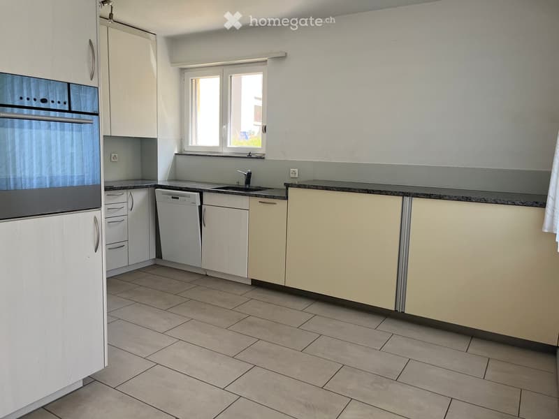 Modern kitchen with white cabinets, tiled floor, and built-in appliances including a dishwasher and oven. Features a window above the sink and a countertop with a backsplash.
