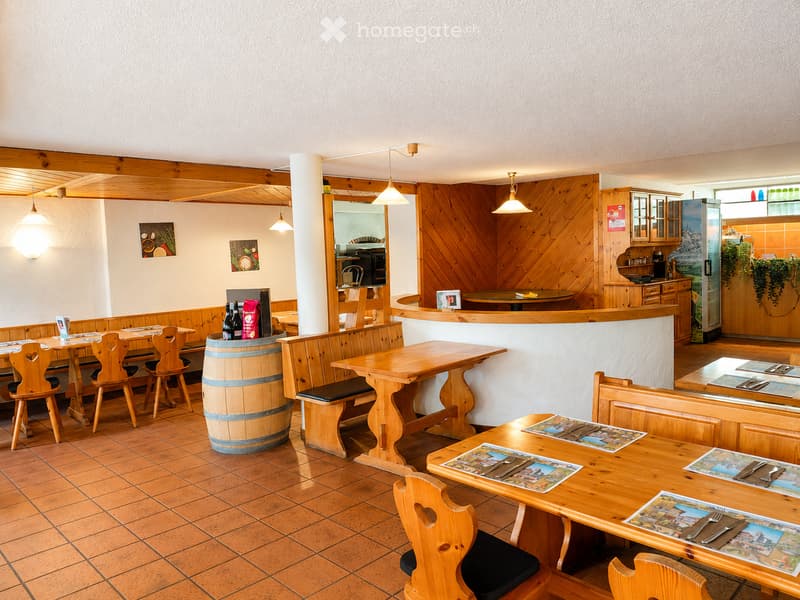Cozy restaurant interior with wooden tables, chairs, benches, and a central barrel. Features wooden paneling, pendant lights, and a bar area with cabinets and a refrigerator.