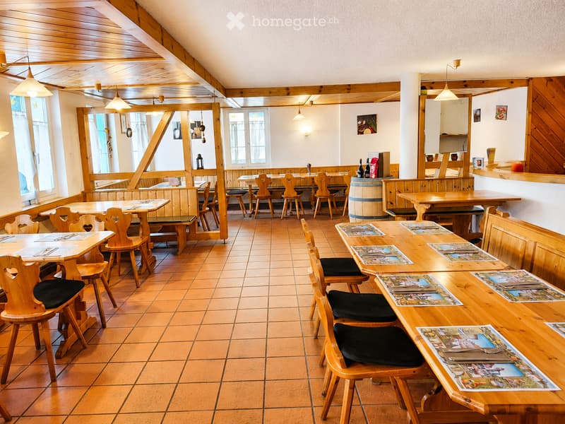 Cozy restaurant interior with wooden tables, chairs, and benches arranged around the space. Menu cards are placed on the tables, and a wooden barrel is visible near the bar area.