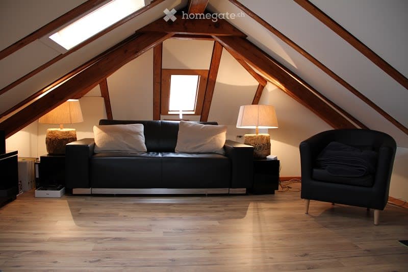 Cozy attic living room with wooden beams, slanted ceilings, and skylights. Features a black leather sofa with white pillows, a matching armchair, side tables, and table lamps.