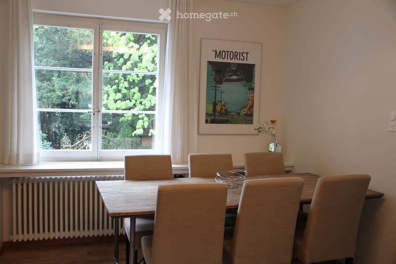 A dining room with a wooden table surrounded by six chairs, a vase with a flower on the table, and a framed picture on the wall. The room has large windows with white curtains and a radiator below them.