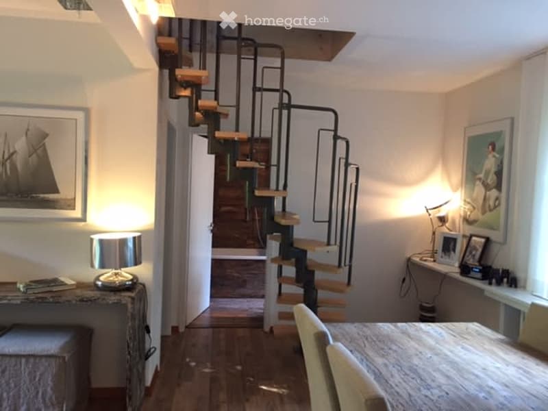 A modern, well-lit room with a wooden floor, a spiral staircase leading upwards, a small table with chairs, a lamp, and picture frames on the walls.