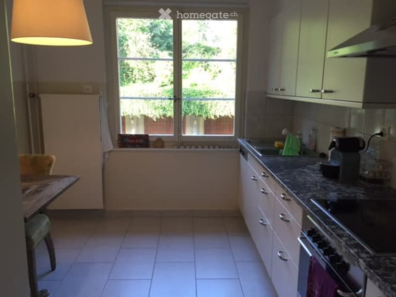 Bright kitchen with white cabinets, granite countertops, and a large window overlooking a garden. Includes a refrigerator, stove, oven, sink, and a small dining area with a table and chair.