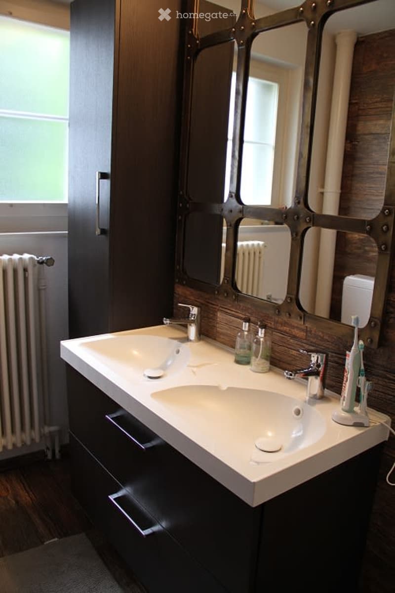 Modern bathroom with a double sink vanity, sleek dark wood cabinetry, and a large mirror with a distinctive geometric frame. The sink features modern chrome faucets, and personal care items are neatly arranged on the counter.