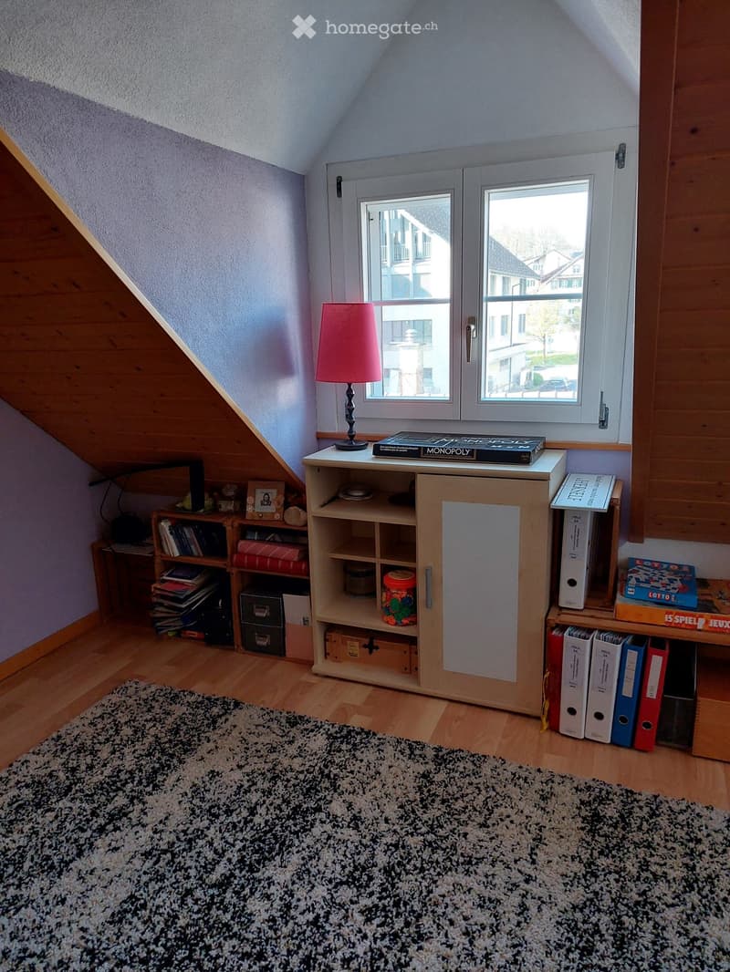 Cozy attic room with wooden floors, slanted ceilings, large window, and various furniture including a table with a lamp, bookshelf, cabinet, and file cabinet.