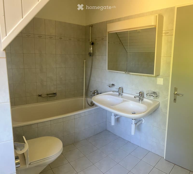 Modern bathroom with a bathtub, sink, toilet, and mirror. The walls are tiled, and the floor is tiled as well.