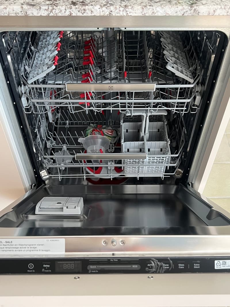 Open stainless steel dishwasher with multiple racks and various dishes inside, including a cup and a bottle.