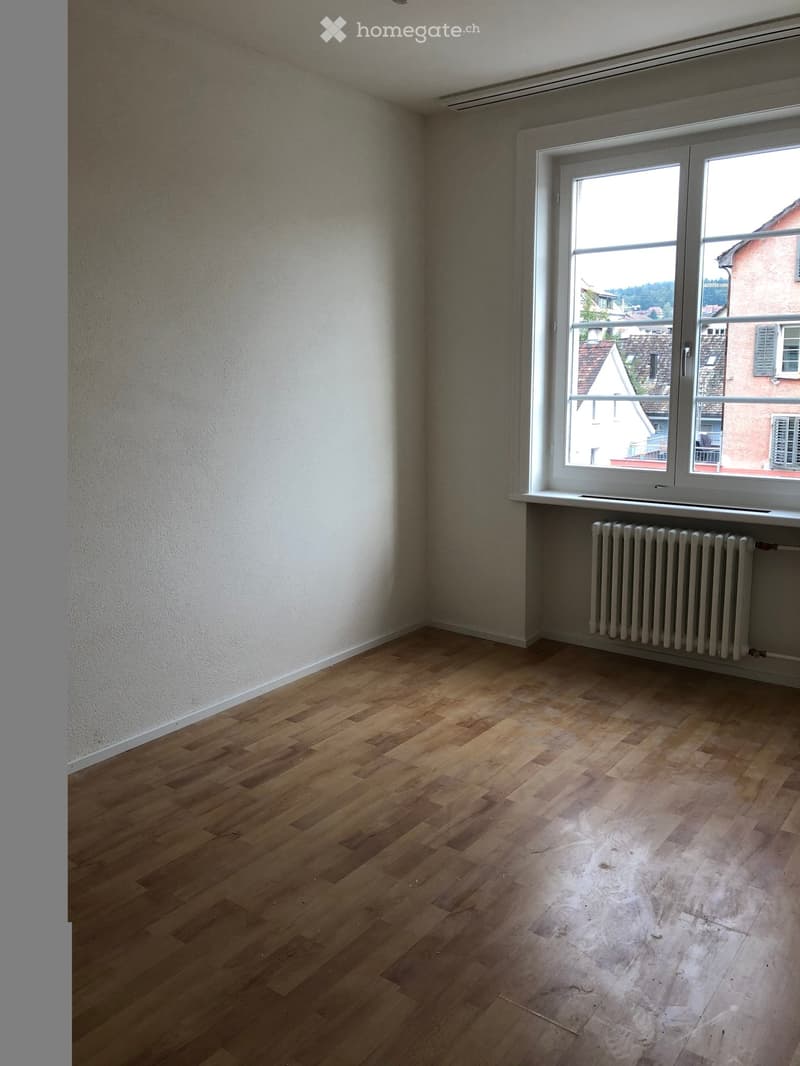 Empty room with wooden flooring, white walls, and a large window with a radiator below it.