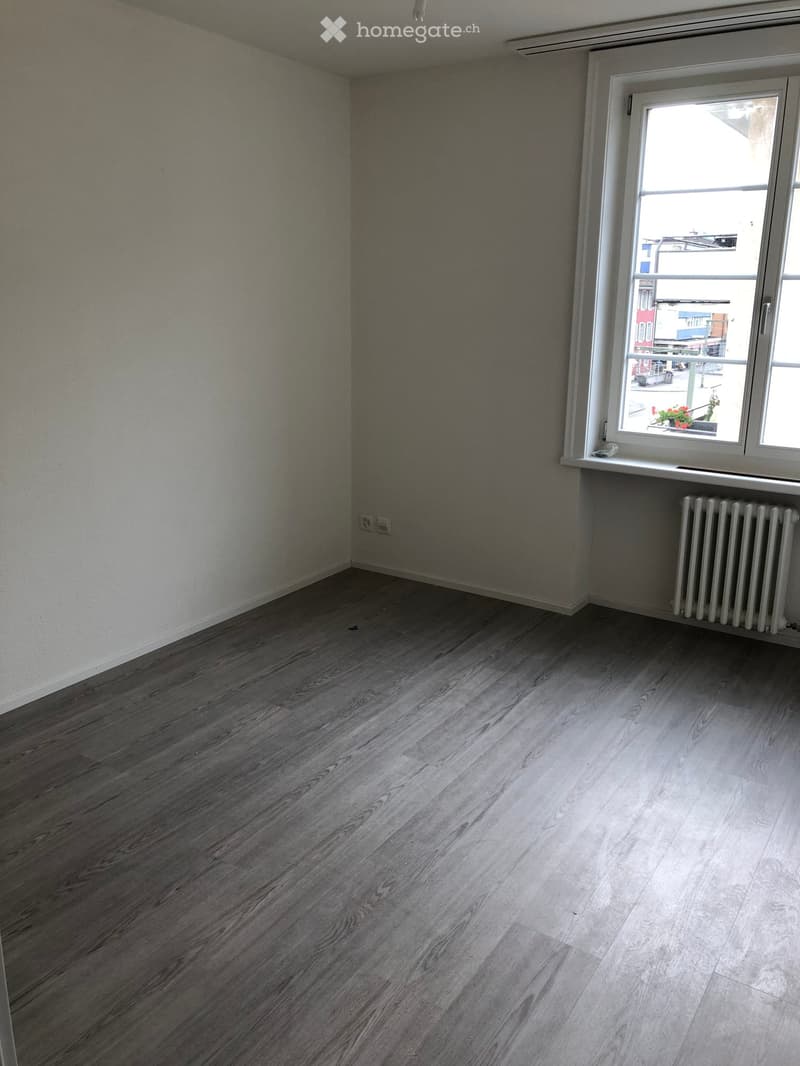 Empty room with wooden flooring, white walls, and a window with a radiator below it.