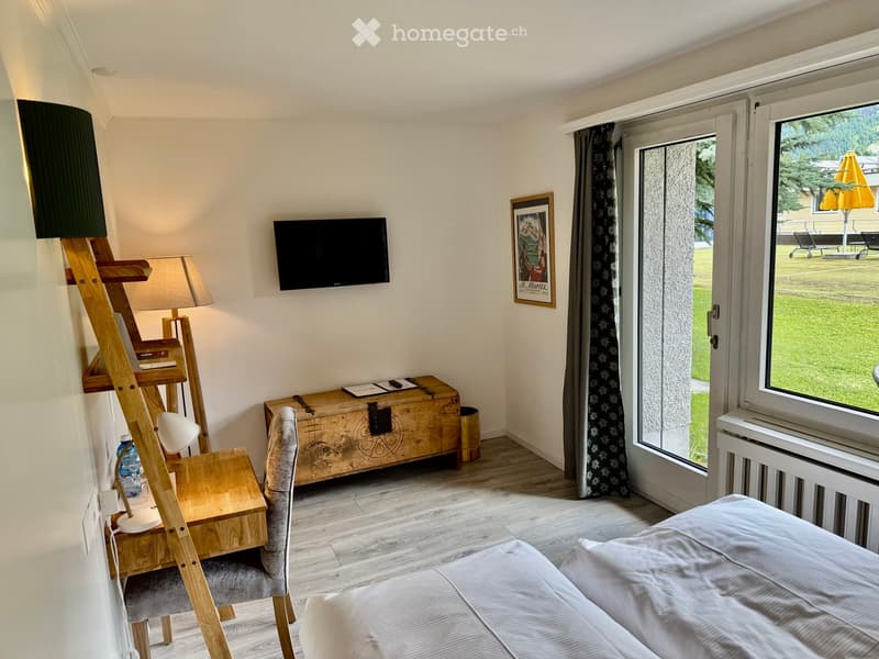 Cozy bedroom with a bed, wooden chair, small table, lamp, television, and picture frame. Large window with view of the garden.