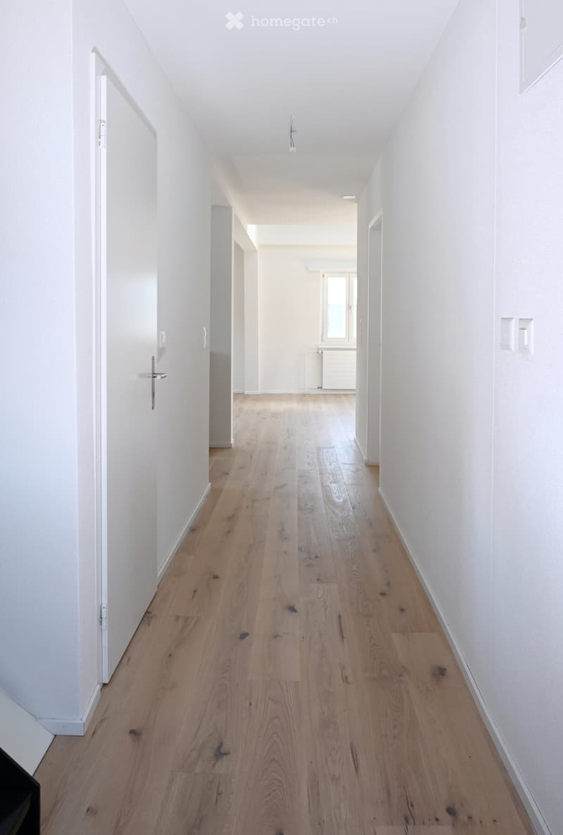 A long, narrow hallway with light wooden flooring and white walls, featuring several doors and a visible room at the end.