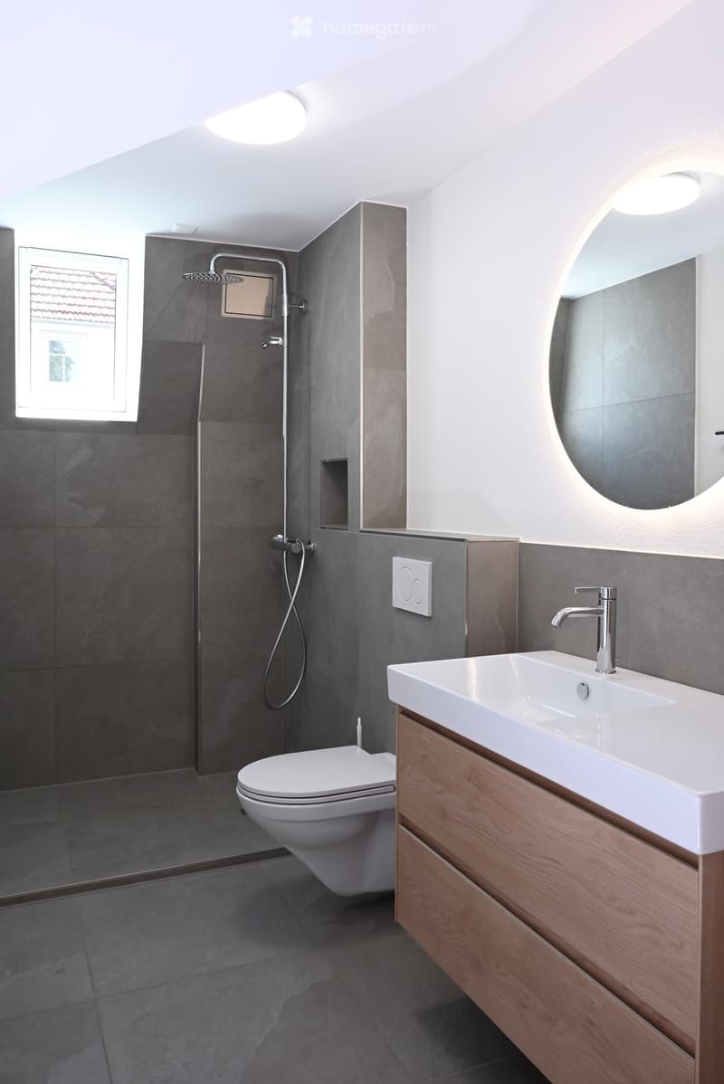 Modern bathroom with dark grey tiles, a white toilet, a white sink with wooden cabinet, a chrome faucet, and a shower with a handheld sprayer.