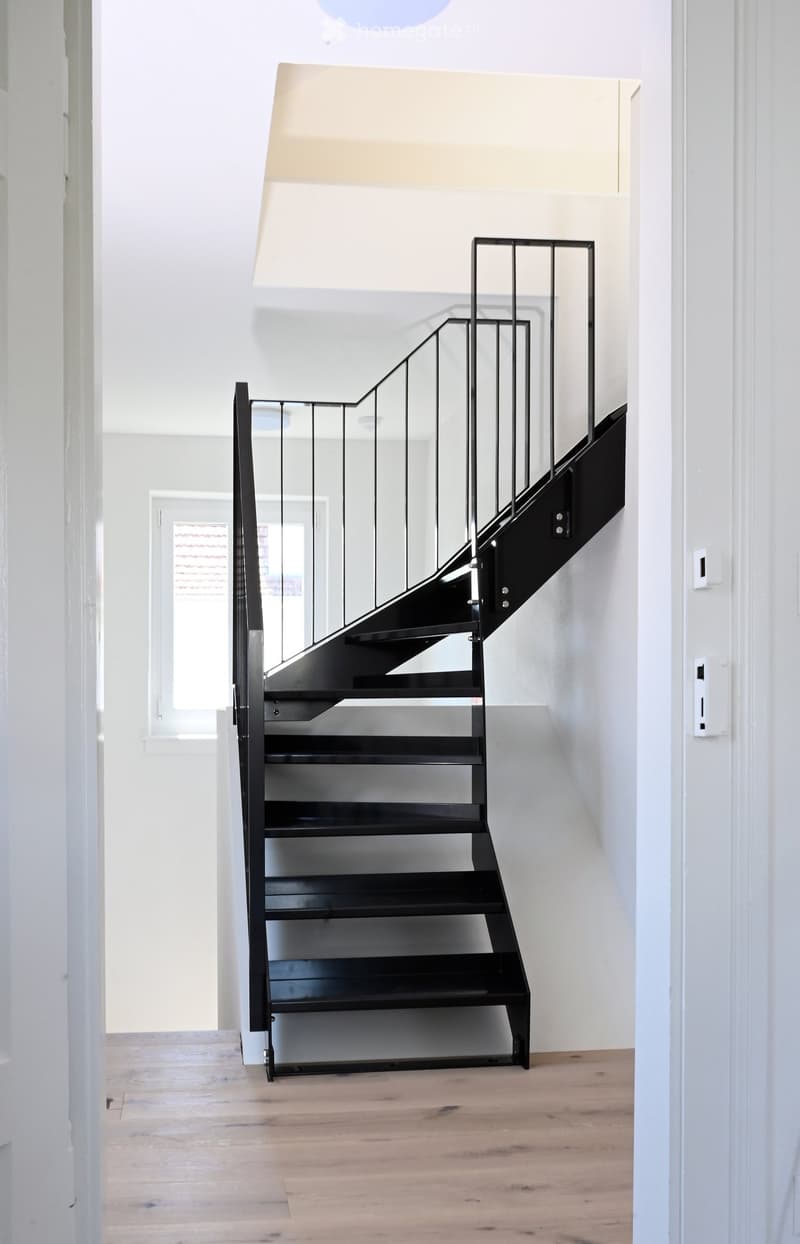 Modern hallway with a sleek black staircase featuring a glass railing, light wooden flooring, and white walls.