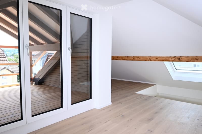 Bright, empty attic room with wooden flooring, white walls, large windows, and a skylight. The room features a sliding glass door leading to a wooden deck with a view of the neighborhood.