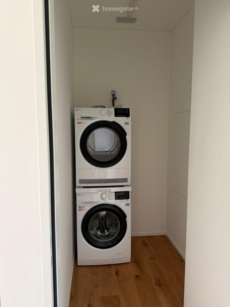 Compact bathroom with a stacked washing machine and dryer unit, light wooden flooring, and white walls.