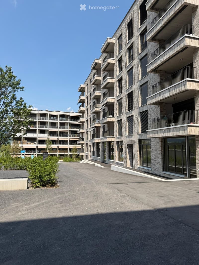Modern multi-story residential building with balconies and large windows, surrounded by greenery and a paved area.