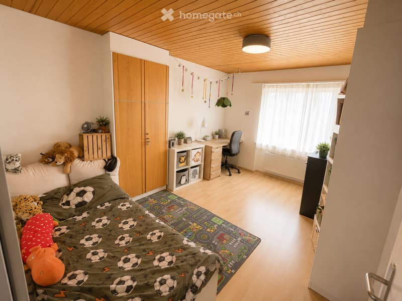 Bright, modern bedroom with wooden flooring and a slanted wooden ceiling. Features a bed with a green soccer-themed duvet, a sofa with stuffed animals, a large wardrobe, a desk with a chair, and a storage unit with books and toys.