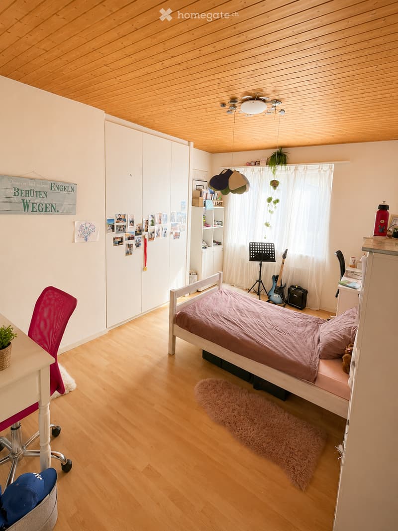 A well-lit bedroom with wooden flooring and a slanted wooden ceiling. Features a single bed with a pink duvet, a red chair, a small table with a plant, a guitar and guitar stand, a mirror, and a white wardrobe with photos and a sign on the wall.