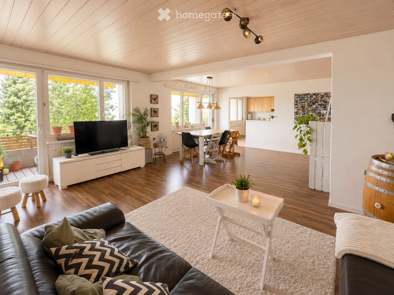 Modern living room with wooden flooring, white walls, and large windows. Features a black leather sofa with patterned cushions, a white coffee table with a candle, a small white stool, a dining area with a table and chairs, and a TV on a white stand. Ther