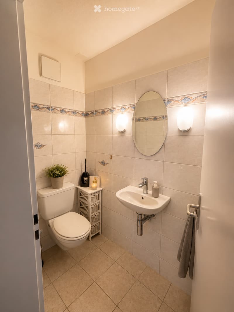 A small, well-lit bathroom with beige tiles, a toilet, a sink with a modern faucet, a round mirror, a towel rack, a small plant, and a storage unit with baskets.