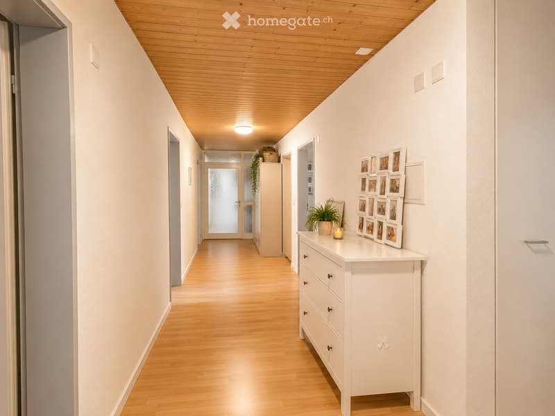 Bright hallway with wooden flooring and white walls, featuring a white dresser with framed photos, a small potted plant, and a candle on top.