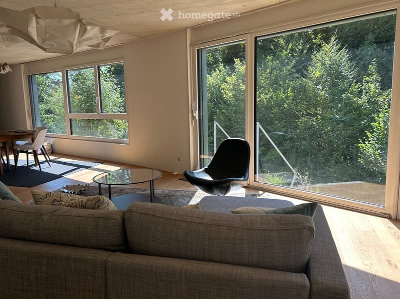 Bright living room with large windows and a view of greenery, featuring a sofa, coffee table, dining area, and modern chairs.