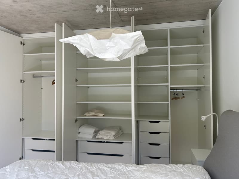 Modern bedroom with a neatly made bed, large white wardrobe with shelves and drawers, and a wall-mounted lamp.