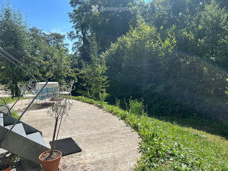 Garden path lined with greenery, small seating area with white metal chairs and a table, potted plant, and a concrete bench on the left side.