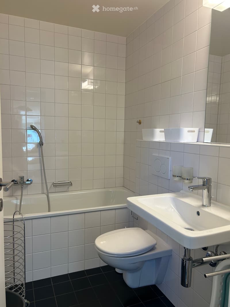 Modern bathroom with white tiled walls, black tiled floor, bathtub, toilet, sink, and showerhead. Includes a mirror, towel rack, and decorative cups on the shelf.