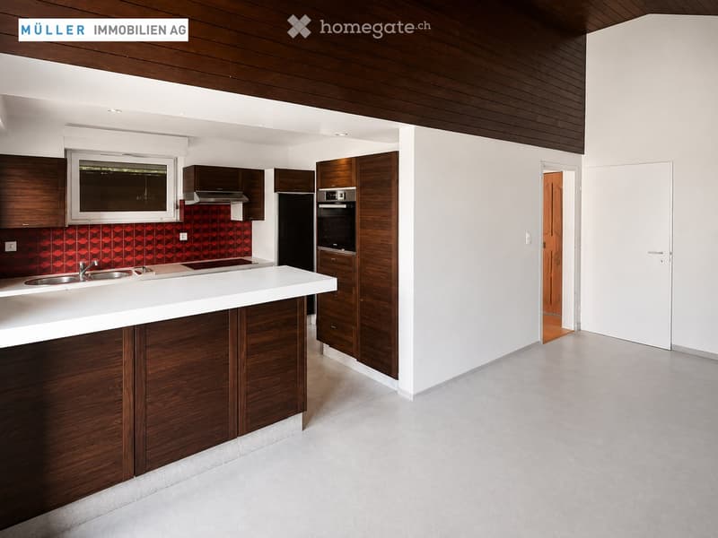 Modern kitchen with white countertops, wooden cabinets, stainless steel sink, and built-in oven. Features a red backsplash and large window.