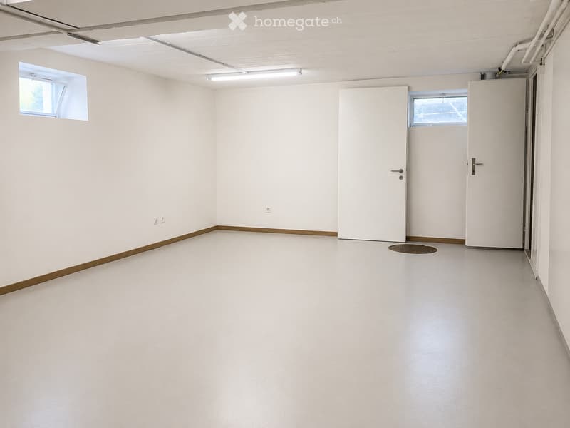 Empty room with white walls, a polished concrete floor, and two doors. There are two small windows on the left wall and a drain cover on the floor.