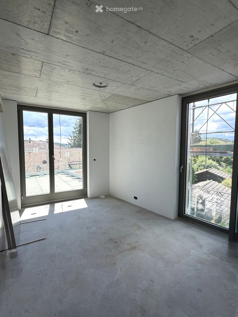Empty room with concrete floors and walls, large windows offering views of rooftops and greenery, exposed wooden beams on the ceiling.
