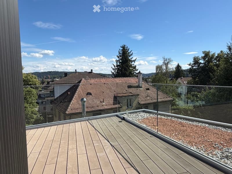 Modern rooftop terrace with wooden decking, glass railing, and a view of the city and surrounding greenery.