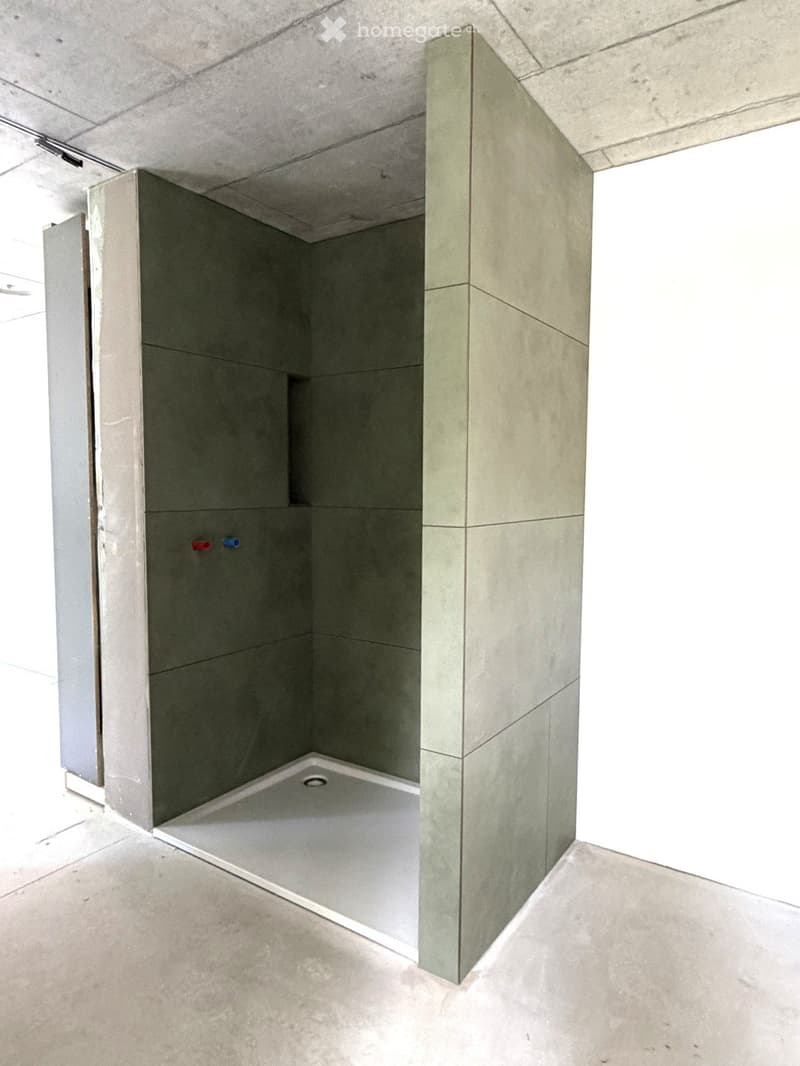 Minimalist bathroom with concrete walls and floor, featuring a single shower stall with a built-in drain and a small niche for toiletries.