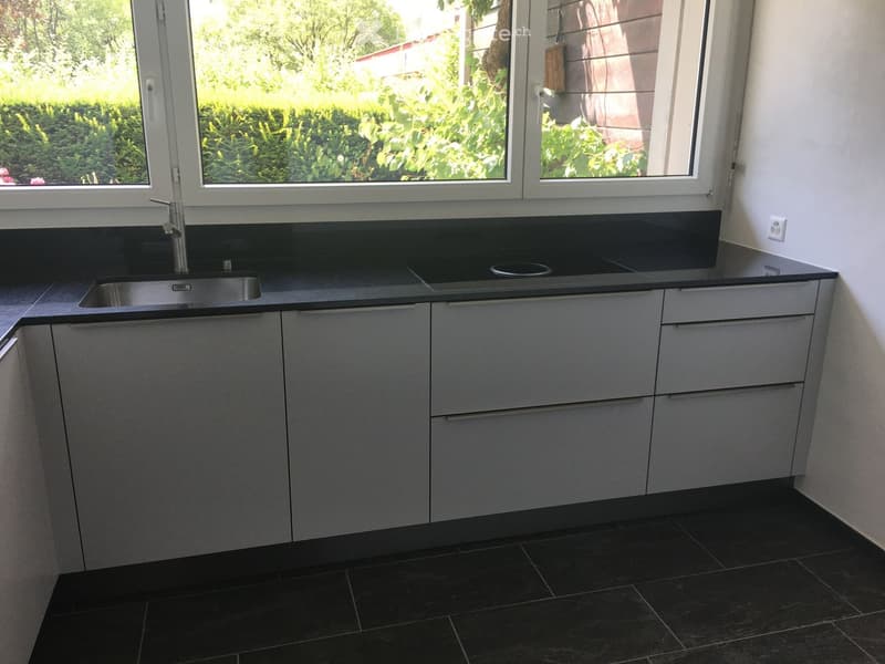 Modern kitchen with grey cabinets, a stainless steel sink, and a black countertop, featuring large windows with a view of greenery.