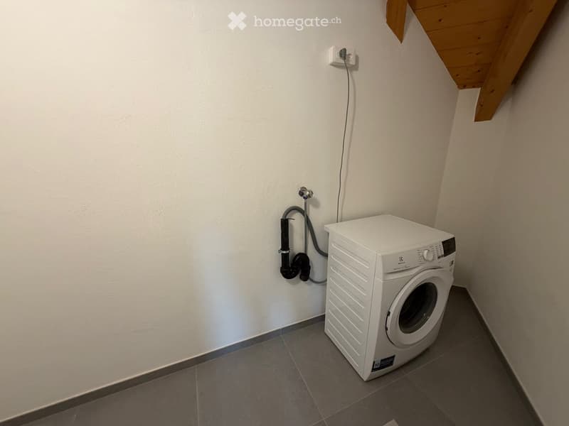 small room, white wall, white washing machine, grey tile floor, plug socket on the wall