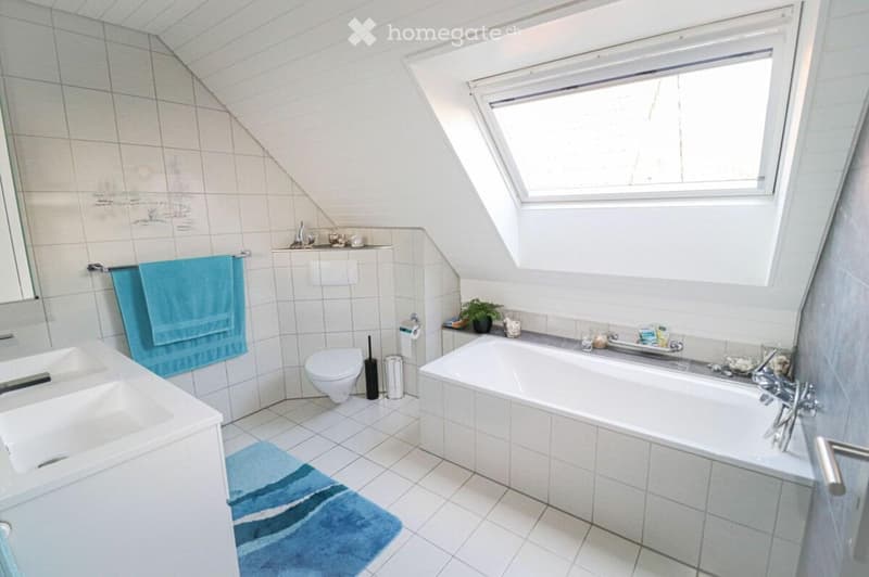 Bright bathroom with a skylight window, featuring a bathtub, toilet, double sink, and blue towels.